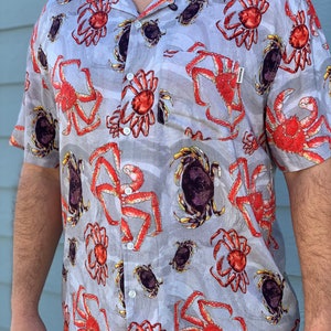 May include: A gray short-sleeved button-down shirt with a repeating pattern of red and black crabs. The shirt has a collar and a button-up front. The brand name "Matthews" is visible on the shirt.