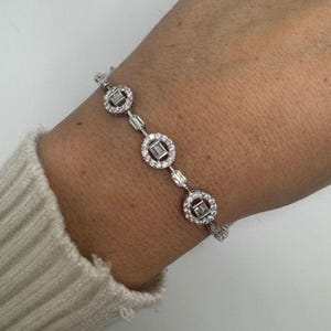 May include: A delicate silver bracelet featuring a series of round and rectangular-cut clear gemstones. The bracelet is composed of multiple circular settings, each encasing a square-cut stone, connected by smaller rectangular stones. The bracelet is worn on a wrist.