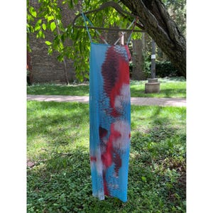 May include: A sleeveless midi dress featuring a vibrant blue base color with a prominent red and black scale-like pattern throughout. The garment is suspended on a hanger and includes thin, crisscrossing shoulder straps at the top neckline.