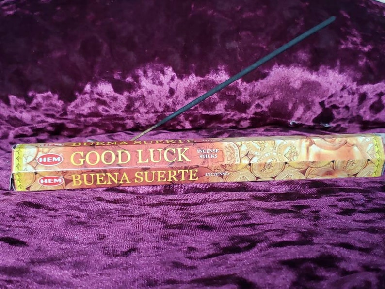 May include: A box of HEM Good Luck incense sticks with a gold and red design. The box has the words "Good Luck" and "Buena Suerte" printed on it. The incense sticks are made with natural ingredients and are designed to bring good fortune and prosperity.