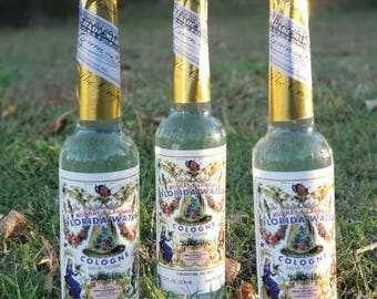 Pode incluir: Três frascos de colônia Florida Water, cada um com uma tampa de folha de ouro e um rótulo com ilustrações florais e botânicas. Os frascos são transparentes, cheios de um líquido verde e são exibidos na grama verde.