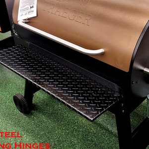 May include: A black powder-coated shelf with a diamond plate pattern. The shelf is attached to a Treager Pro Series 34 grill with stainless steel folding hinges. The grill is brown and has a black handle.