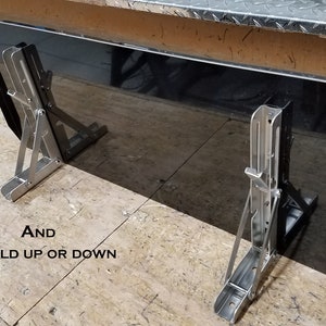 May include: A black rectangular panel with two stainless steel folding brackets. The brackets are attached to the panel and can be folded up or down. The text "BRACKETS ARE STAINLESS STEEL" is visible in the image. The text "joecarparts" is also visible in the image.