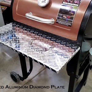 May include: A silver folding shelf made with highly polished aluminum diamond plate. The shelf is attached to a black and brown smoker grill. The text "FITS PIT BOSS TAILGATER" is visible on the left side of the image.