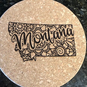 May include: A cork trivet with a black and white design of the state of Montana with the word "Montana" written in a decorative font.
