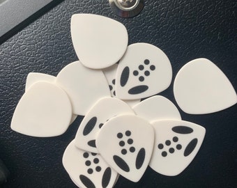 Può includere: Un mucchio di plettri per chitarra bianchi con disegni neri. Alcuni plettri hanno un semplice motivo a punti, mentre altri hanno un design più complesso con due ovali neri e un gruppo di punti.