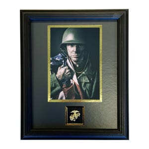 May include: Framed print featuring a soldier in a military uniform holding an American flag. The image is set within a black frame with a gold border. A small gold emblem is displayed below the main image.
