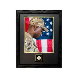May include: A framed photo of a soldier in military uniform standing in front of an American flag. The soldier is wearing a camouflage hat and is looking down. A gold coin is displayed below the photo.