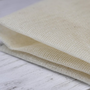 Op de afbeelding: Close-up van een gevouwen, gebroken witte, geweven stof. De stof heeft een fijne, open weefselstructuur en de randen zijn netjes gevouwen. De textuur lijkt zacht en enigszins doorschijnend. De achtergrond is een lichtgekleurd houten oppervlak.