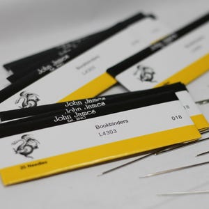 May include: A collection of John James bookbinding needles and packaging. The packaging is white with black and yellow accents, featuring the brand name and logo. The needles are silver and scattered around the packaging.