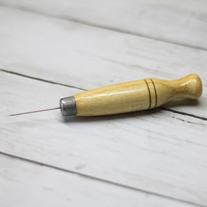 May include: A wooden awl with a metal tip and a light brown handle. The handle has two dark brown bands near the top. The awl is lying on a white wooden surface.
