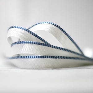 May include: White ribbon with a blue stitched border, arranged in graceful loops. The ribbon's texture is visible, and the blue stitching provides a contrasting detail. The image is set against a soft, blurred background.