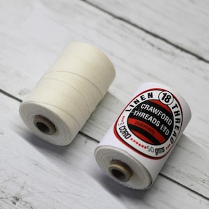 May include: Two spools of linen thread. One is a solid off-white color. The other has a label that reads "Linen Threads Ltd" with the number "18" and "50 gms". The spools are on a light wood grain surface.