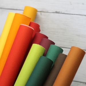 May include: A collection of rolled felt sheets in various colors, including yellow, orange, red, green, and brown. The felt rolls are arranged in a fan shape, showcasing the different hues and textures of the material. The background is a light-colored wooden surface.