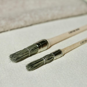 May include: Two paint brushes with light wooden handles and silver ferrules. The bristles are a dark green colour. The text "A BETTER HAIR" is printed on the handles. The brushes are on a white surface.