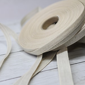 Puede incluir: Primer plano de varios rollos de cinta beige con diseño de espiga. La cinta está enrollada en carretes, con una parte desenrollada y plana. La cinta tiene una textura sutil y un color neutro, adecuada para varios proyectos de manualidades.