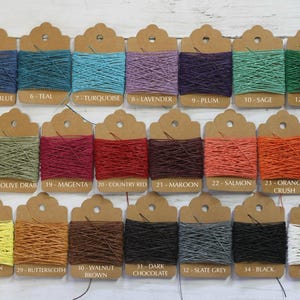 Pode incluir: Uma coleção de rolos de barbante coloridos exibidos em etiquetas marrons. Os rolos são de várias cores, incluindo azul, verde, roxo e laranja. Cada etiqueta tem um número e um nome de cor, como "Azul Royal" e "Teal".