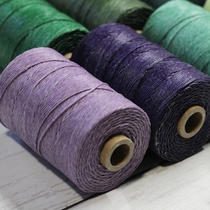 Puede incluir: Primer plano de varios carretes de hilo de lino encerado de colores. Los carretes son en tonos morado, verde y turquesa. El hilo está bien enrollado alrededor de un núcleo de cartón. La imagen está tomada sobre una superficie de madera blanca.