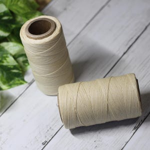 Puede incluir: Dos carretes de hilo beige, uno en posición vertical y otro de lado, sobre un fondo de vetas de madera blancas y grises. El hilo está bien enrollado en núcleos de cartón. Se ven plantas verdes con hojas en el fondo.