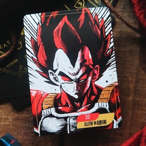 May include: A trading card featuring a black and red illustration of a character with spiky black hair and red eyes. The character is wearing a white and red outfit with gold accents. The card has the text "2/12 Alien Warrior" in the bottom right corner.
