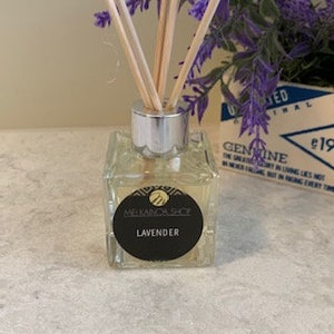 May include: A clear, square glass diffuser bottle with a silver cap and wooden reeds. The bottle has a black label that reads "Lavender" and "Melanida Shop." Purple lavender sprigs are in the background, along with a white box with blue text.