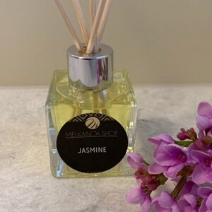 May include: A clear glass bottle diffuser with a silver cap and wooden sticks. The bottle is filled with a yellow liquid and has a black label that reads "MELGAINOA SHOP JASMINE".