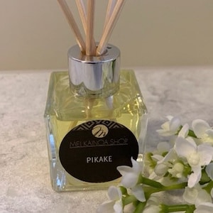 May include: A clear glass bottle with a silver lid and wooden sticks, filled with a yellow liquid. The bottle has a black label with the text "Mei Kainoa Shop" and "Pikake".