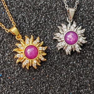 May include: Two sun-shaped pendant necklaces. One is gold-toned with a pink center stone and clear accent stones. The other is silver-toned with a similar pink center stone and clear accent stones. Both are on delicate chains.