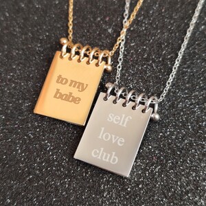 May include: Two pendant necklaces. One is gold-toned with the words "to my babe" engraved on a rectangular pendant. The other is silver-toned with "self love club" engraved on a similar pendant. Both have a spiral binding design.