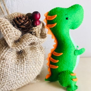 May include: A green felt dinosaur toy with orange stripes and black eyes. The dinosaur is standing next to a hessian sack.