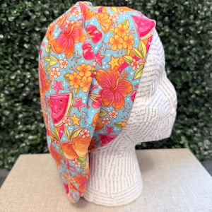 May include: A scrub cap featuring a light blue background with a vibrant pattern of pink hibiscus flowers, orange citrus slices, red cherries, watermelon wedges, and small yellow stars. The fabric is gathered and worn on a white mannequin head.