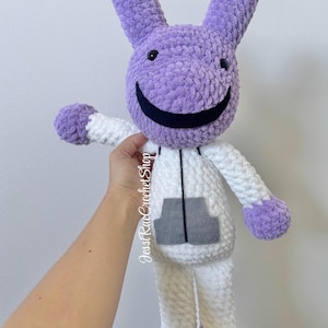May include: A plush stuffed animal with a purple head, long ears, and a wide black smile. The body is white with purple hands and feet, featuring a gray rectangular pocket on the front. The text JesstRaeCrochetShop is visible on the side.