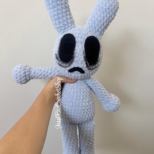 May include: A light blue crochet plush toy shaped like a rabbit with long ears, large black oval eyes, and a small black frown. The plush has a textured, yarn-based surface and features the text JessRaeCrochetShop printed vertically on its side.