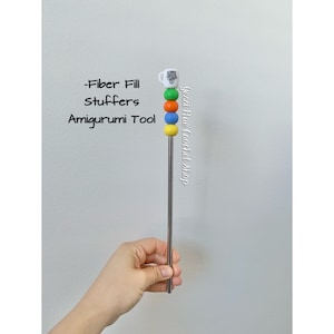 May include: An amigurumi tool with a stainless steel shaft and colorful accents. The tool features a white mug charm and a series of colored beads: green, orange, blue, and yellow. Text on the tool reads "-Fiber Fill Stuffers Amigurumi Tool".