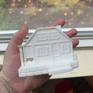 May include: A white 3D printed model of a small house with a porch and a chimney. The house has windows and a door. There are logs stacked on the porch.