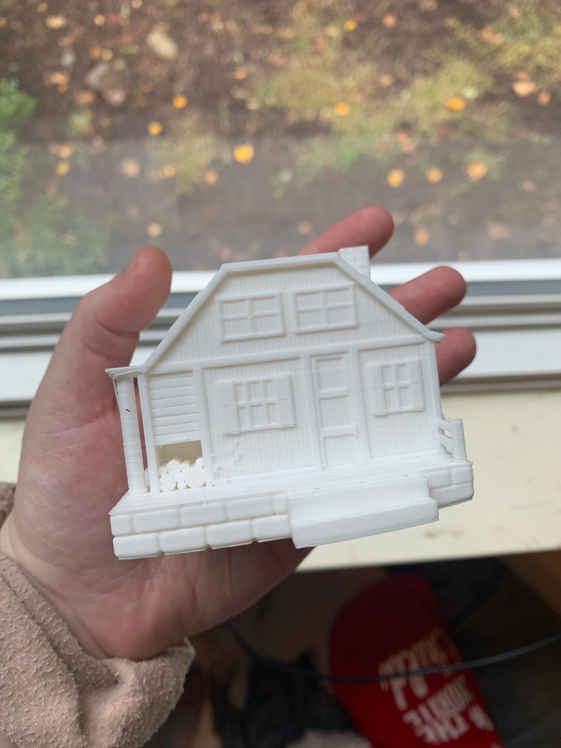 May include: A white 3D printed model of a small house with a porch and a chimney. The house has windows and a door. There are logs stacked on the porch.