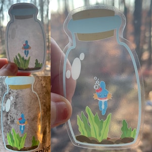 May include: A clear glass jar with a blue lid, containing a blue and white cartoon character with a red heart on its chest. The character is surrounded by green seaweed and bubbles. The jar is on a white background.