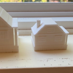 May include: Three white 3D printed model houses of different sizes. The houses are all simple in design with a pitched roof and a single window on each side.