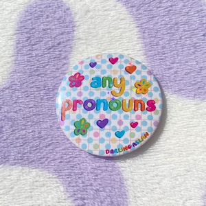 May include: A white button with a blue, white, and pink polka dot background. The button says "any pronouns" in rainbow letters with colorful flowers and hearts surrounding the text. The bottom of the button says "Darling Aslan".