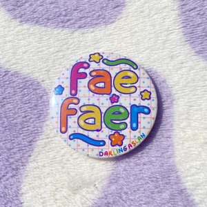 May include: A white button with a rainbow colored graphic that says "fae faer" with a white grid pattern and stars. The button also has the text "DARLING ASLAN" in a blue font.