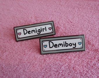 Könnte beinhalten: Zwei Emaille-Pins mit silbernen Rändern und weißem Hintergrund. Der obere Pin sagt "Demigirl" mit einem rosa Herzen auf jeder Seite. Der untere Pin sagt "Demiboy" mit einem blauen Herzen auf jeder Seite.