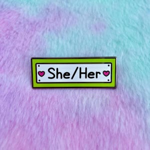 May include: Green enamel pin with a white background and silver border. The pin says "She/Her" with a pink heart on each side of the text.