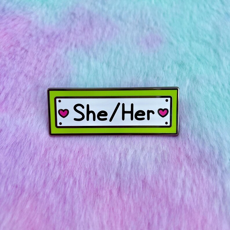 May include: Green enamel pin with a white background and silver border. The pin says "She/Her" with a pink heart on each side of the text.