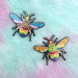 May include: Two rainbow enamel pins shaped like bees with hearts on their wings. The bees have a yellow head, black eyes, and a rainbow striped abdomen.
