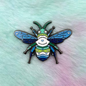 Pu&ograve; includere: Uno spillo in smalto blu, verde e bianco a forma di ape con un corpo peloso e ali glitterate. L'ape ha un viso a forma di cuore con cuori bianchi sul suo corpo.