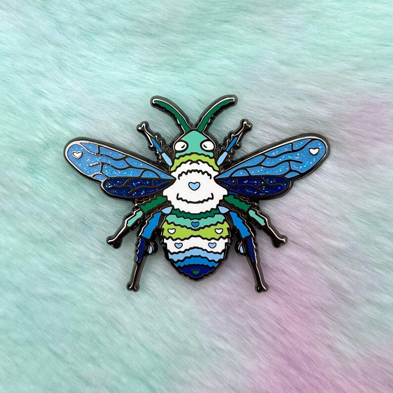 Pu&ograve; includere: Uno spillo in smalto blu, verde e bianco a forma di ape con un corpo peloso e ali glitterate. L'ape ha un viso a forma di cuore con cuori bianchi sul suo corpo.