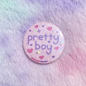 May include: Pink and purple button with white polka dots and hearts. The button says "pretty boy" in purple letters. The text "Darling Aslan" is printed in white on the bottom of the button.