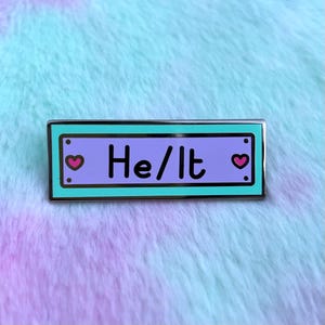May include: A teal blue enamel pin with a purple background and white text that reads "He/It". There are two small pink hearts on either side of the text.