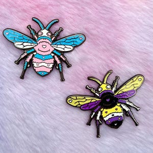 May include: Two enamel pins shaped like bees. The first bee is blue, pink, and white with a heart pattern. The second bee is yellow, purple, and black with a heart pattern.