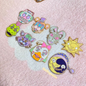 May include: A set of eight enamel pins featuring cartoon characters with colorful details. The characters include a bear, a rabbit, a sun, a moon, a chicken, a frog, and two robots. The pins are all gold-toned and have a glossy finish.